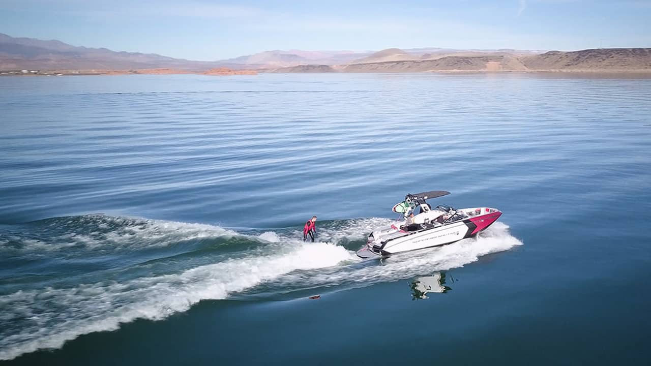 Speed boat driving on a lakebed with a water surfer following behind on a wave.
